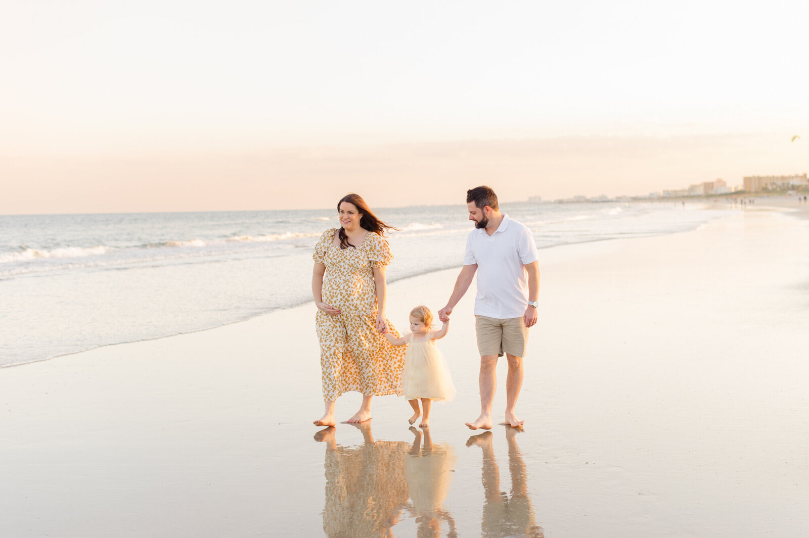 Pregnant mom holds hands with husband and daughter near the shoreline at golden hour