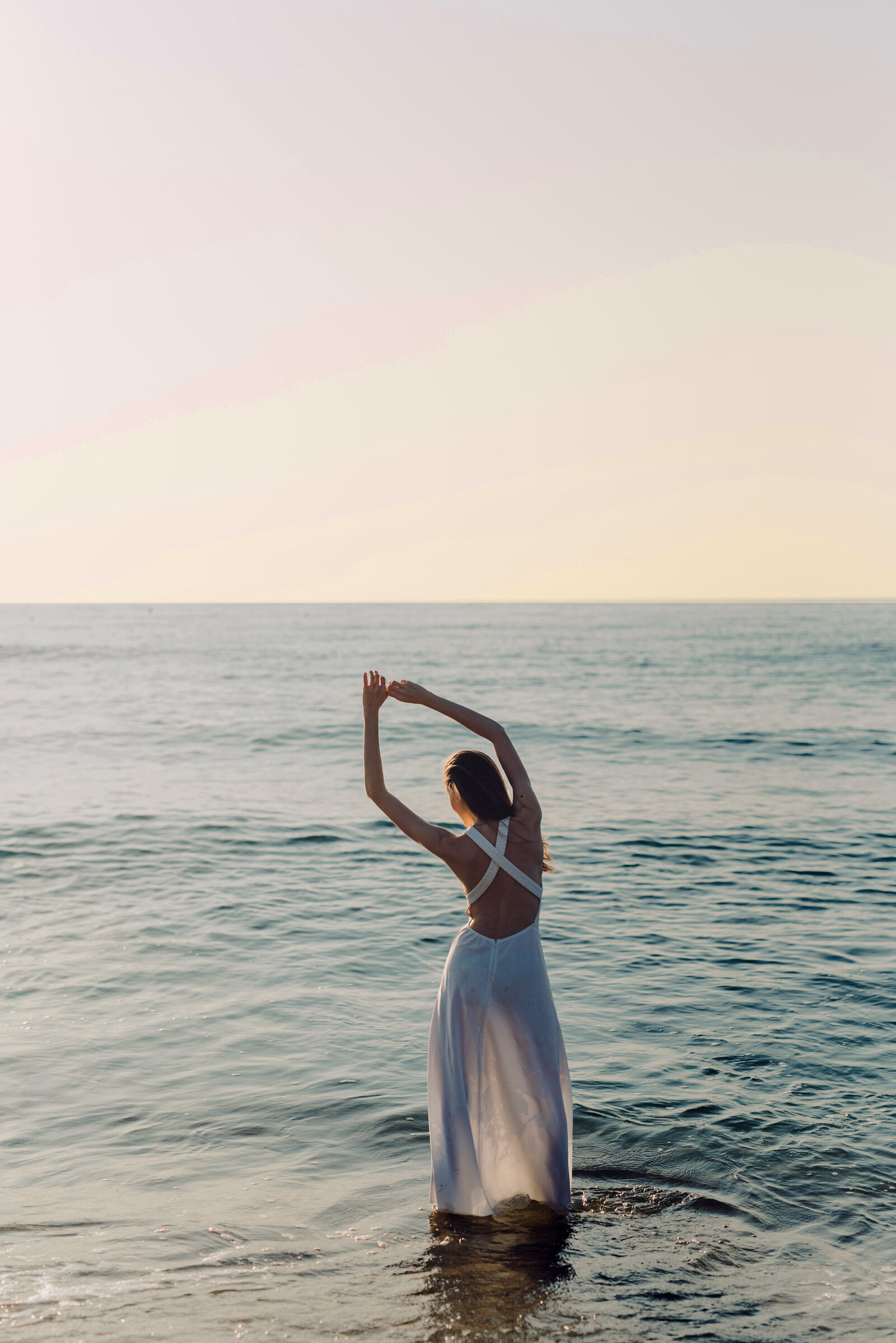 Femme bras levés, vêtue d'une longue robe blanche, de dos, les pieds dans l'eau.