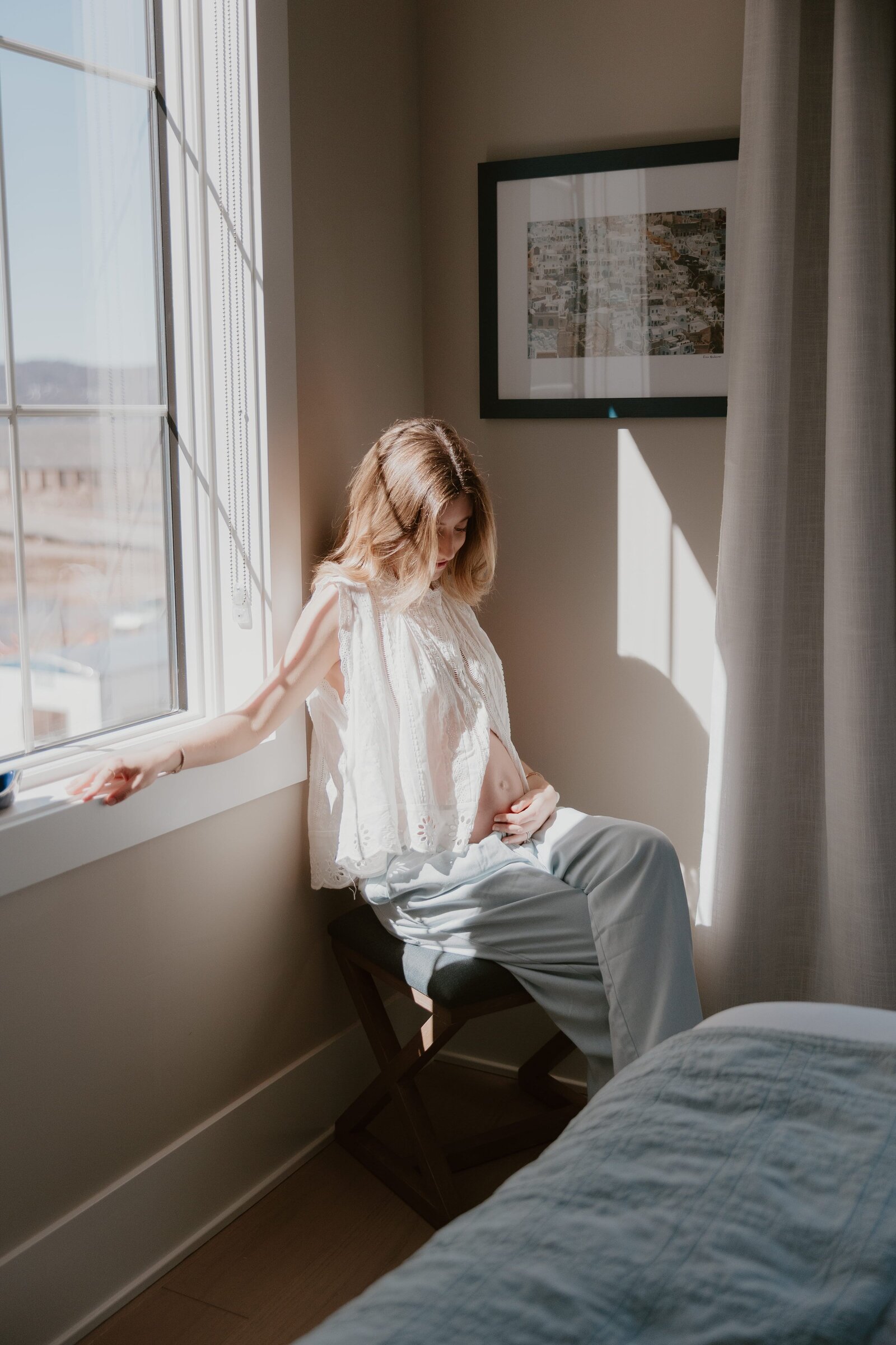 A pregnant woman sits quietly on a stool by a sunlit window, gently cradling her growing belly. She wears a loose white eyelet top and soft blue pants, bathed in warm natural light. The intimate moment feels peaceful and reflective, capturing the beauty of early motherhood in a cozy, light-filled bedroom.