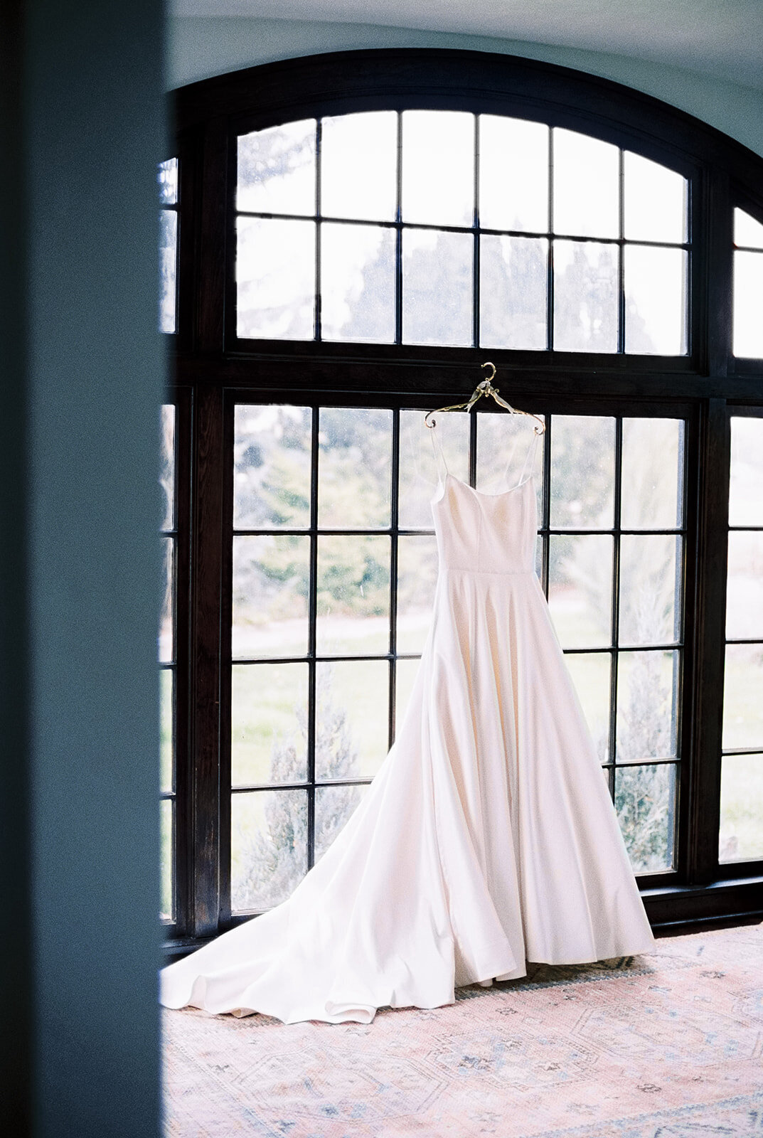 Wedding dress hanging on antique gold hanger on windows at Cottage at Riverbend Wedding by the Best Boise Wedding Photographers