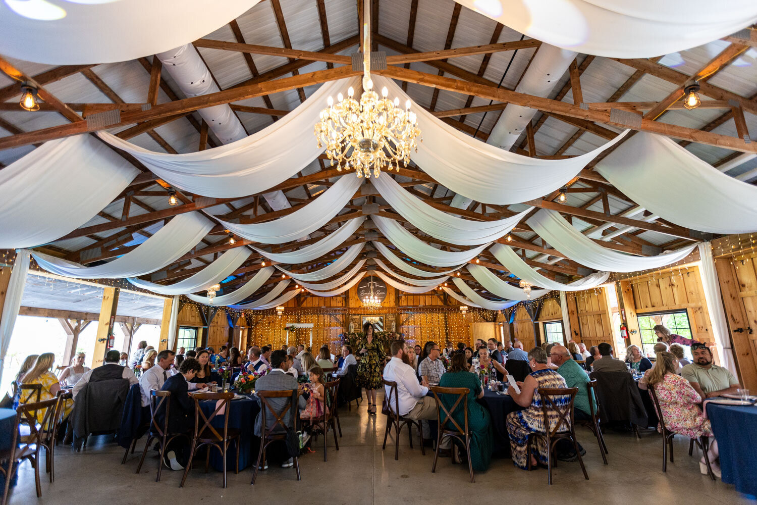Reception in a rustic barn with draped ceiling fabric and chandelier lighting