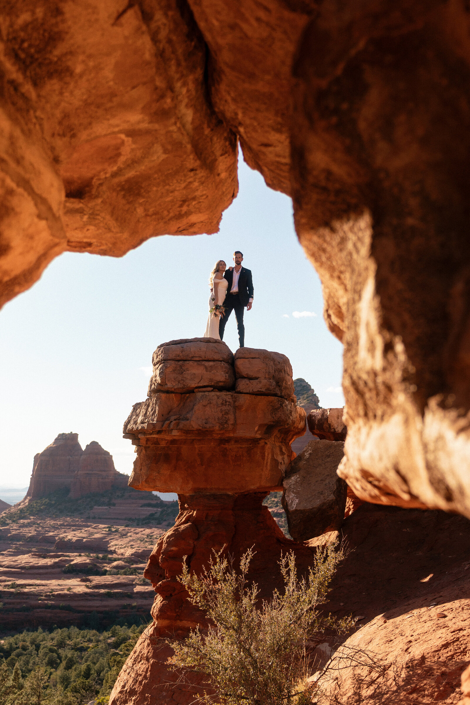 Bride and groom standing on a towering red rock pedestal at Merry Go Round Rock in Sedona, Arizona, framed by natural rock formations with scenic desert views and Cathedral Rock in the background.