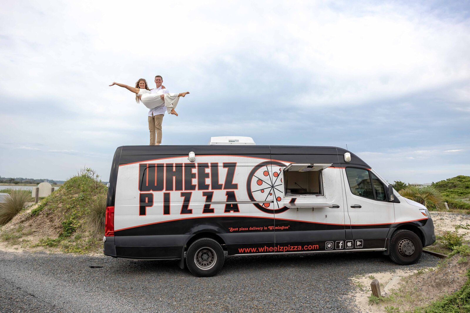 The bride and groom celebrate atop a pizza truck from Wheelz Pizza at a Ocean Isle Beach, NC wedding, showcasing a fun and unique wedding day moment with coastal charm.