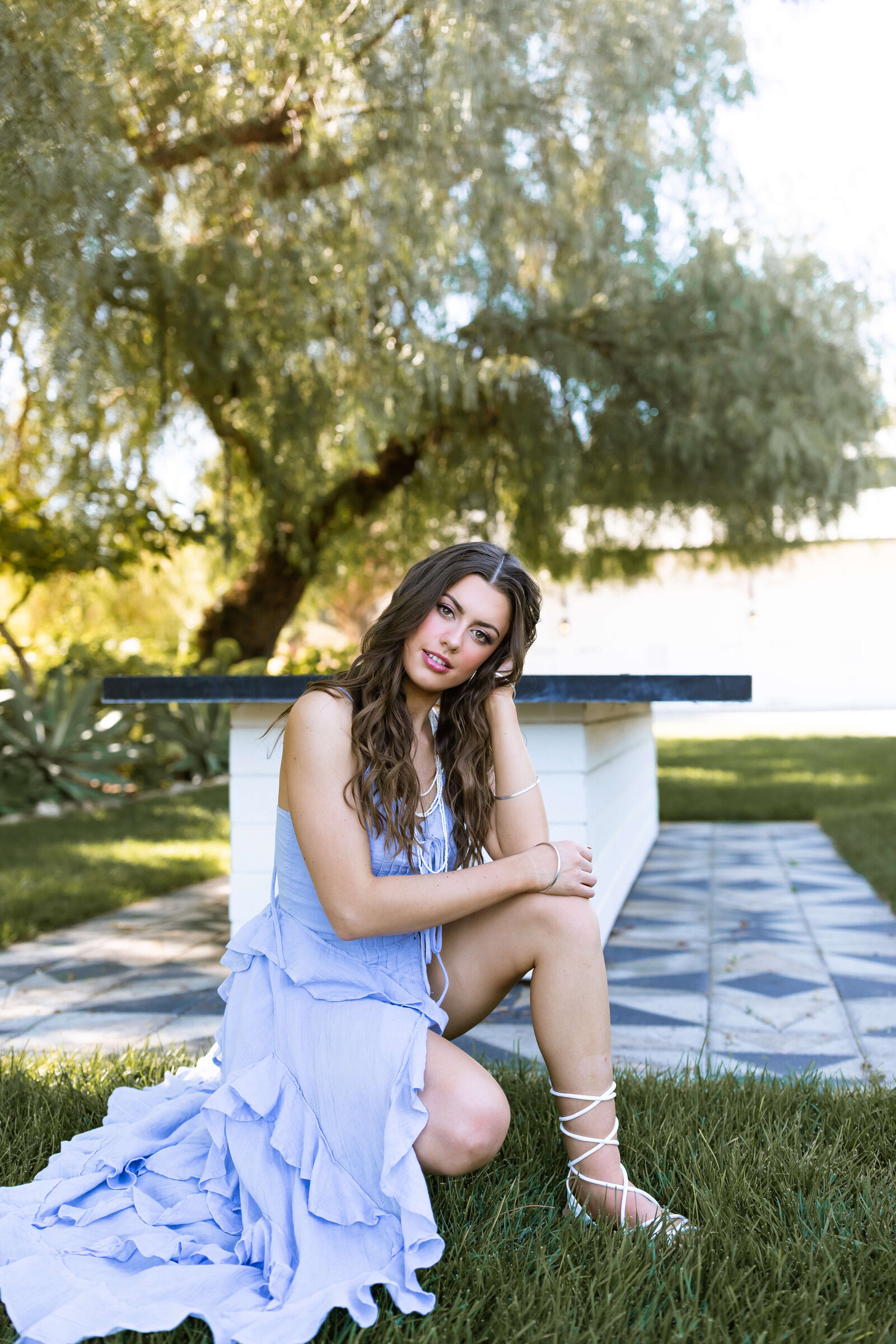 brunette wearing strappy heels and blue dress with pearl necklace standing in front of a table posing for the camera