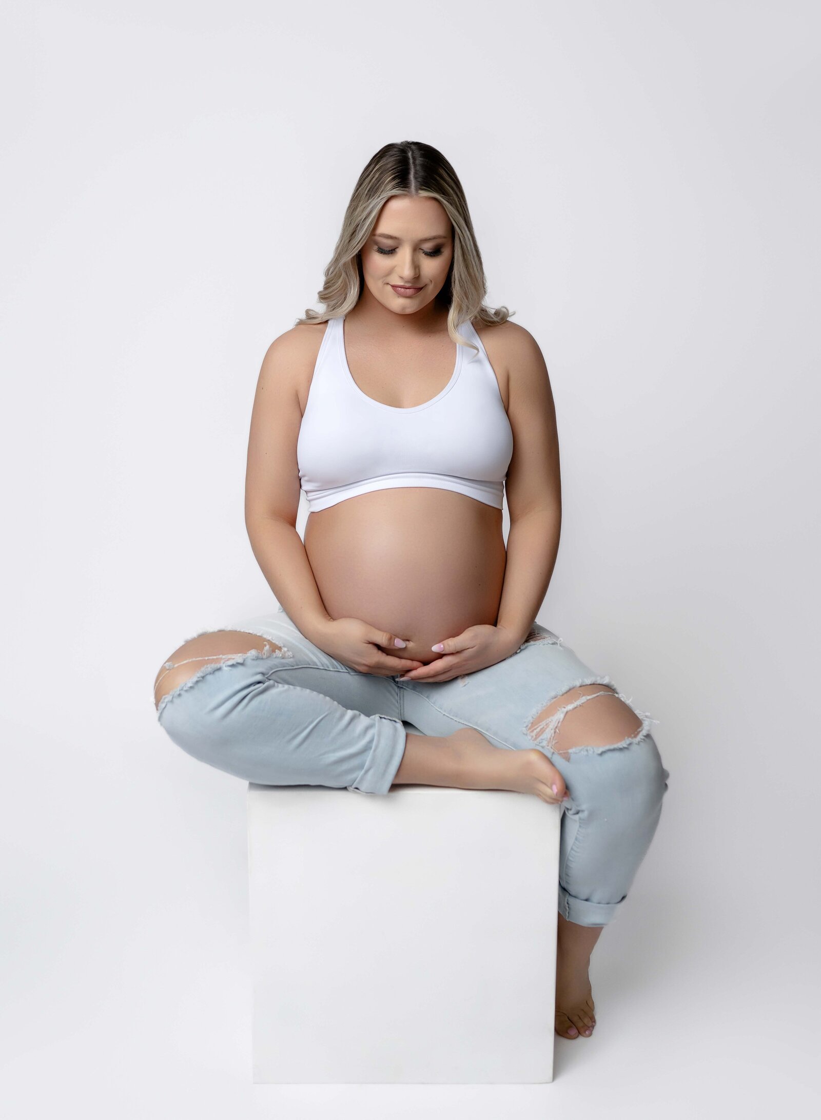 Mom sitting on white box and holding baby bump. Maternity portrait taken by Jenni. Nikki Photography at Atlanta Photo Studio. 