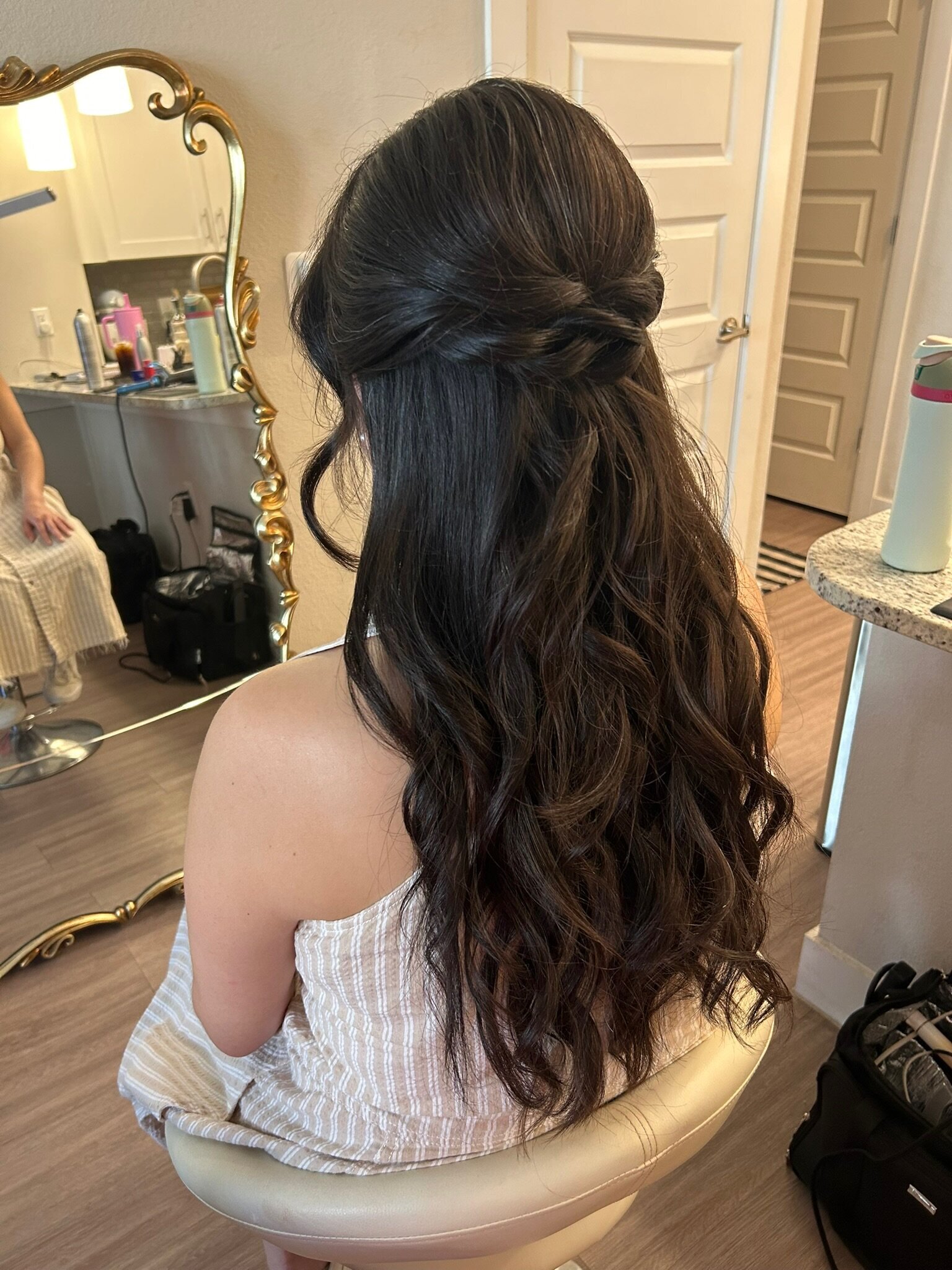 A woman with a long, wavy, half-up hairstyle sits in front of a decorative mirror in a softly lit room, epitomizing effortless wedding hairstyles.
