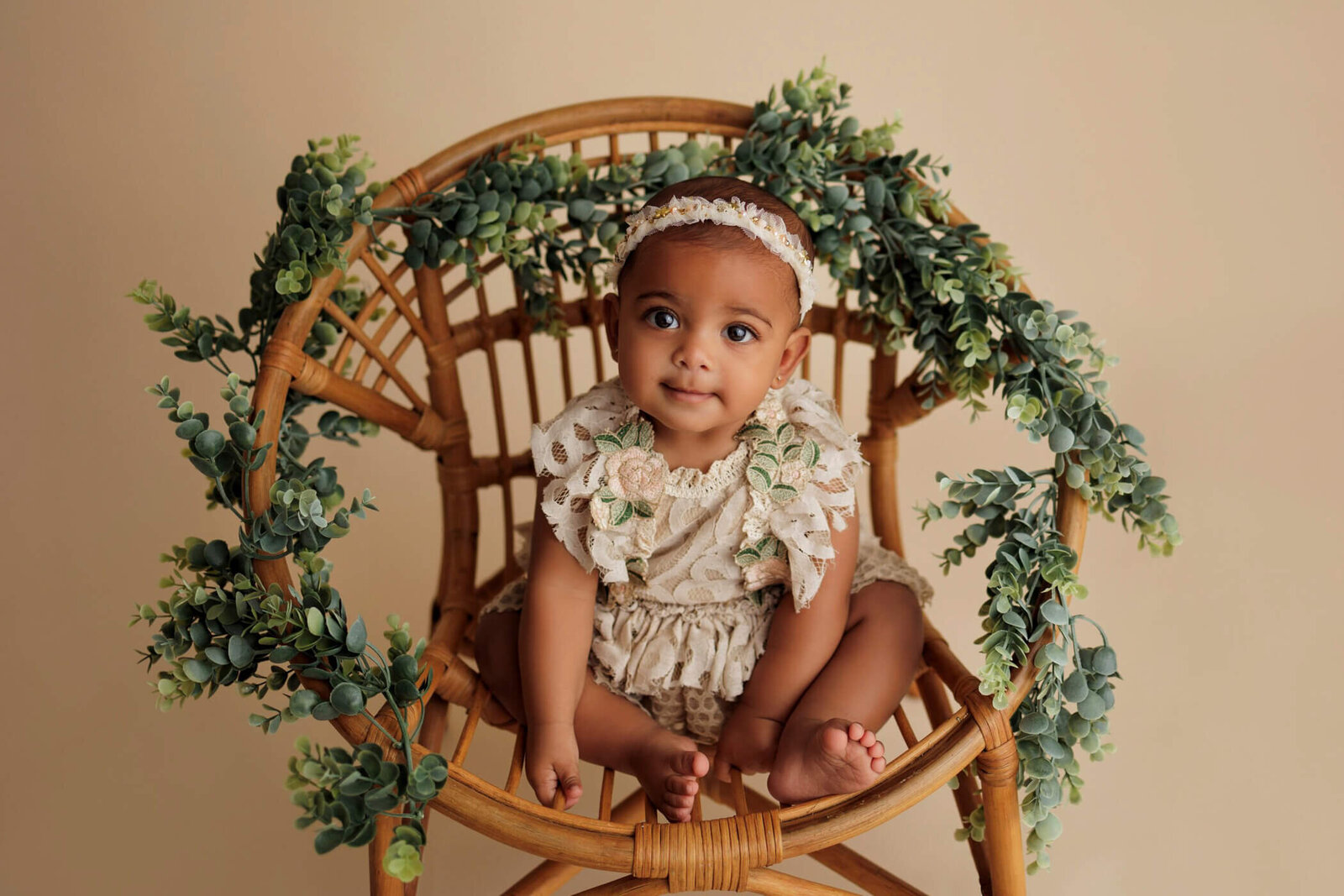 An adorable baby with dark skin and bright eyes sits in a wicker chair adorned with green foliage. The baby wears a cream-colored lace romper and a matching headband with floral accents, looking up with a sweet and curious expression. The background is a soft beige, enhancing the natural and serene atmosphere of the scene.