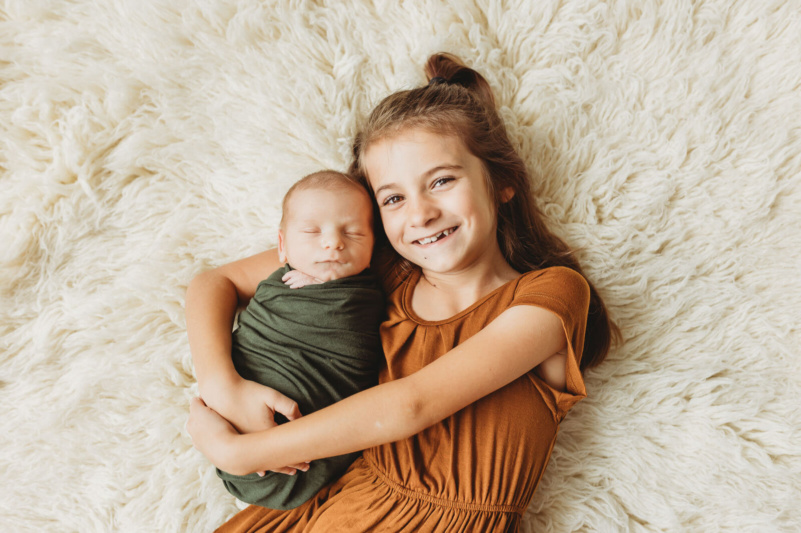 Big sibling holding baby brother who is swaddled laying on a fuzzy rug for newborn photos
