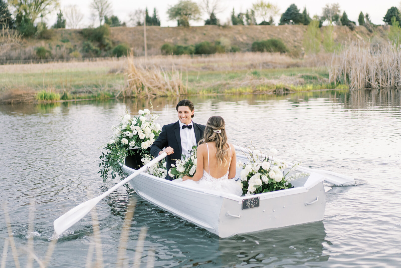 Portrait of a bride and groom wearing a white wedding gown and black tuxedo sitting in a boat filled with flowers on a lake.