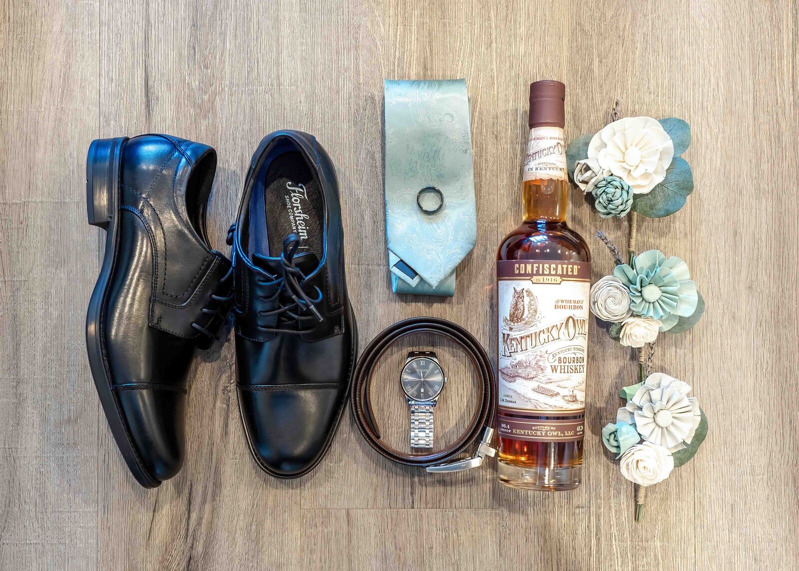A flat lay of groom’s accessories, including polished black shoes, a blue tie, a belt, a watch, and a bottle of Cape Fear whiskey, accented with rustic floral details, perfect for a Wrightsville Beach, NC wedding.