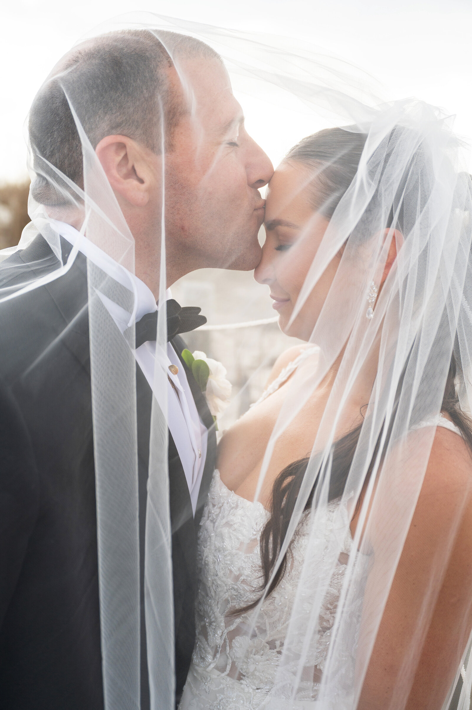 Groom kissing his bride on the forehead while both are under a veil