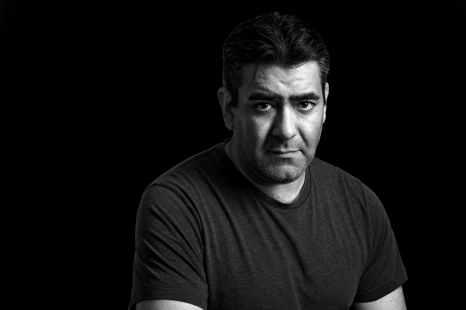 Black and white portrait of a middle-aged man with short dark hair, wearing a plain t-shirt, looking directly at the camera with a serious expression against a solid black background—ideal for music photography Jacksonville.