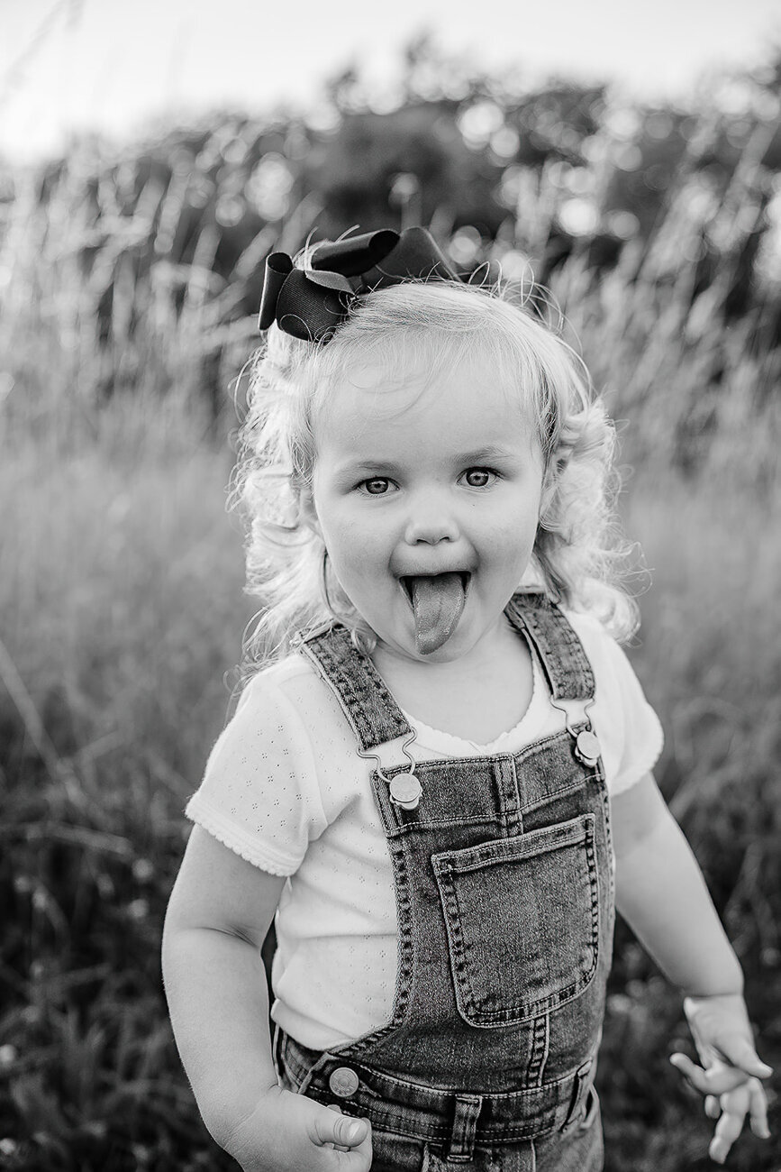 black and white portrait of toddler girl in overalls sticking out tongue