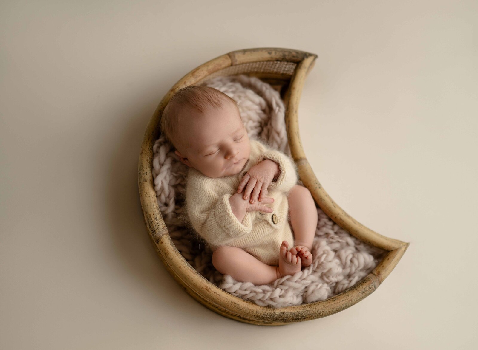 Newborn baby boy in moon boy in moon bowl with cream outfit and hand folded on chest. Photographed at Atlanta newborn photo studio.