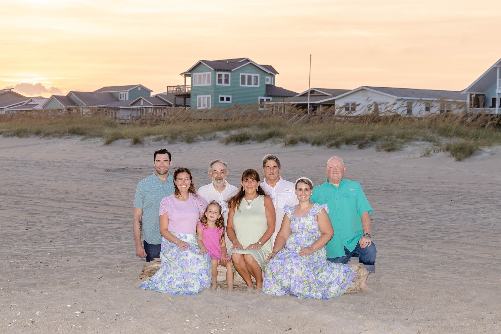 A family poses on the beach at sunset, with coastal homes and sea oats in the background. Dressed in pastel and floral outfits, they capture a serene moment, perfect for North Carolina family photography, highlighting beachside living.