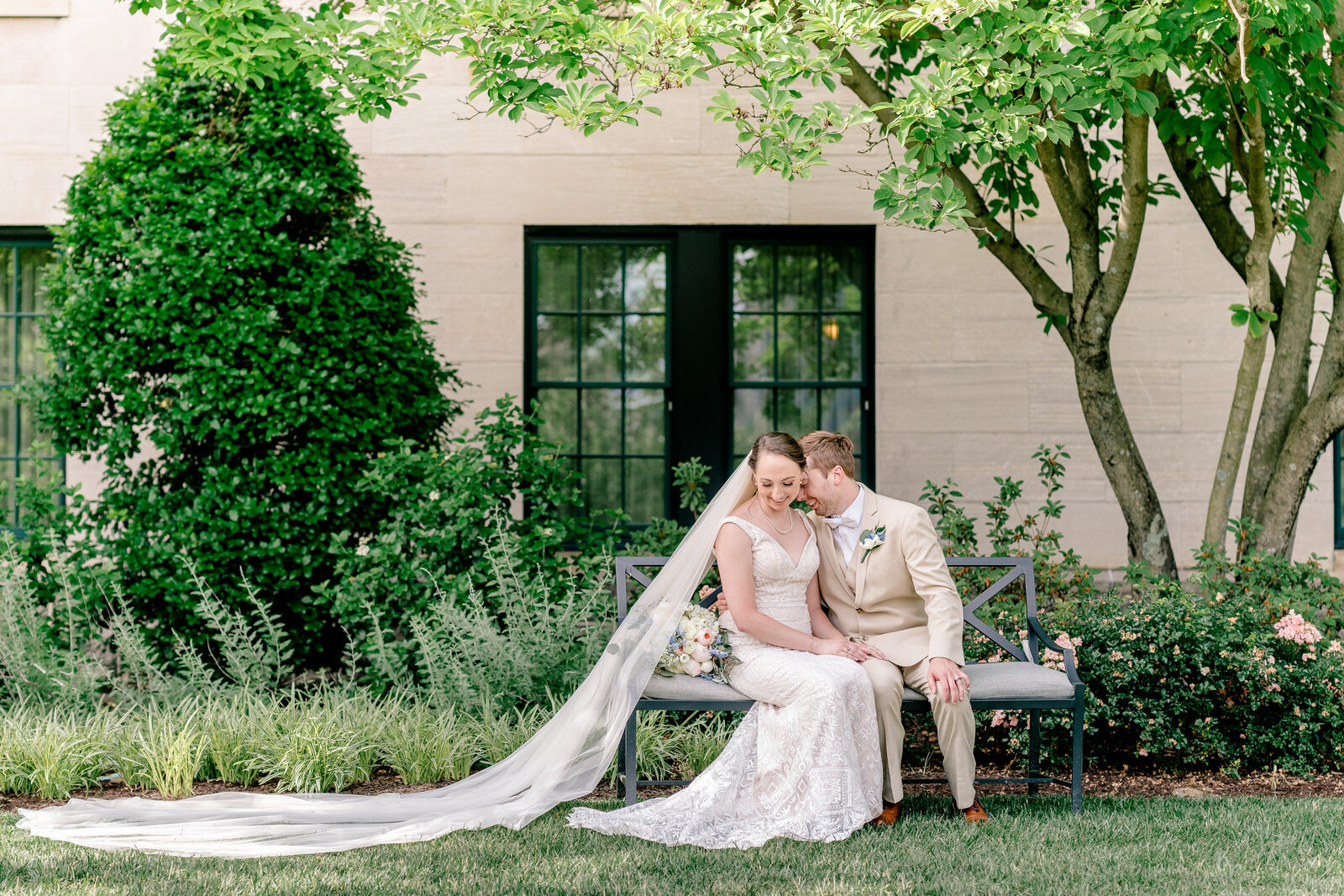 A bride and groom sit on a bench in a garden with her cathedral length veil trailing off camera during their wedding in Northern Virginia