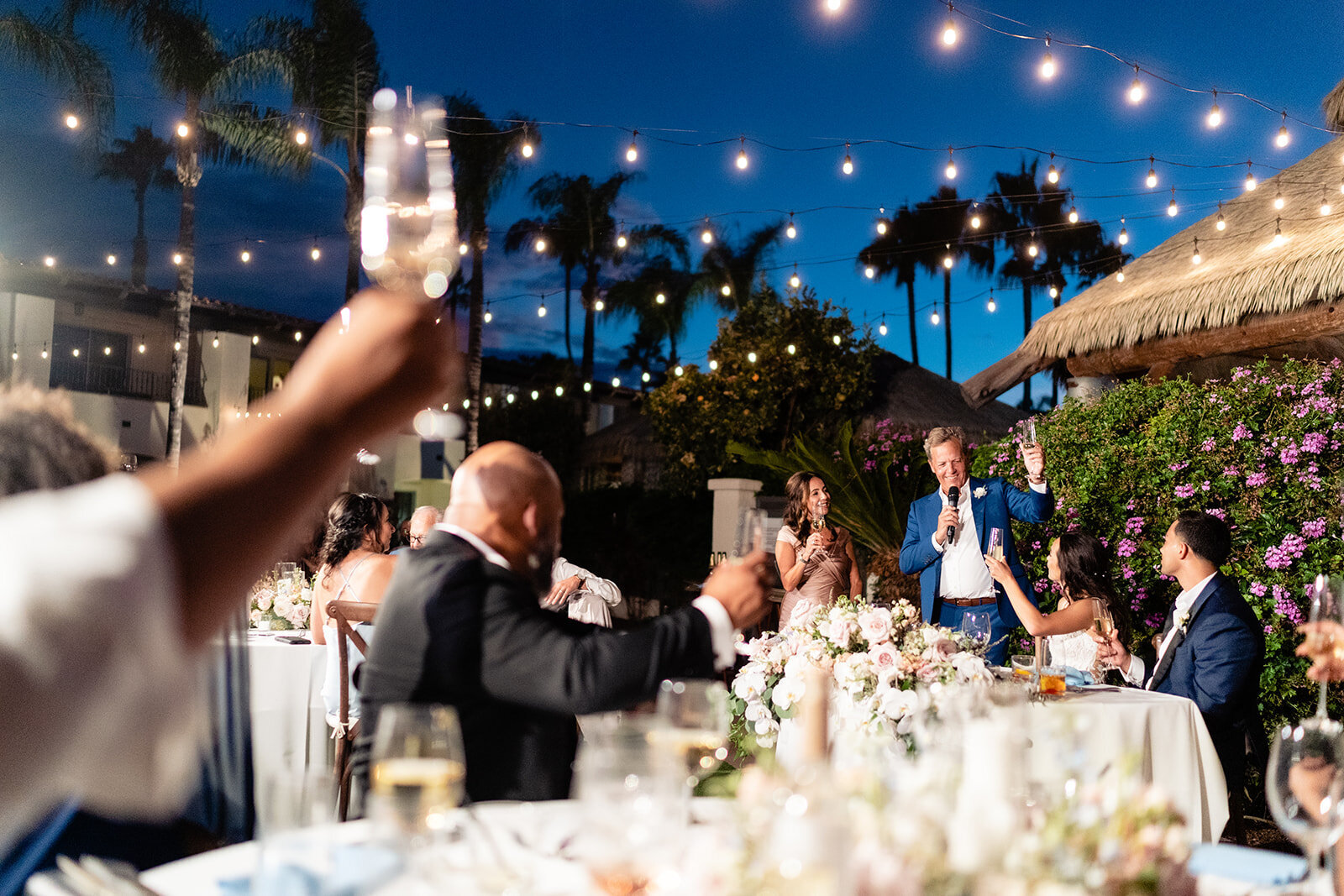 The Father of the groom is giving a toast to the couple while their guests raise their drinks with string lights above and a beautiful dark blue sky at Kona Kai