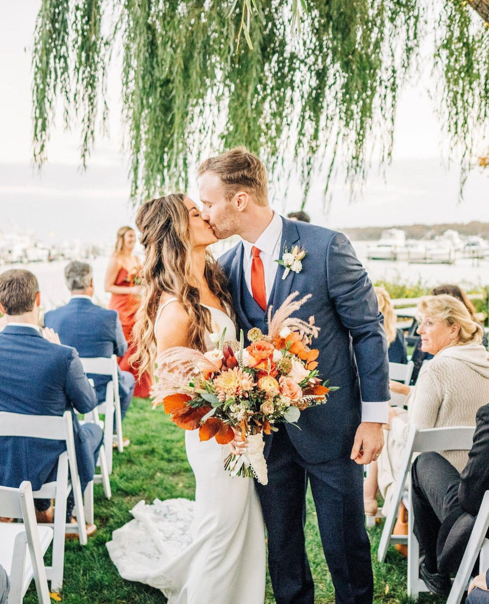 bride holding a colorful bouquet and her groom kissing during their outdoor wedding ceremony and guests are seated in white chairs on a lush green lawn