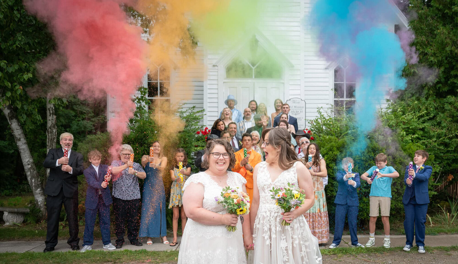 brides laugh together in front of rainbow smoke cannons