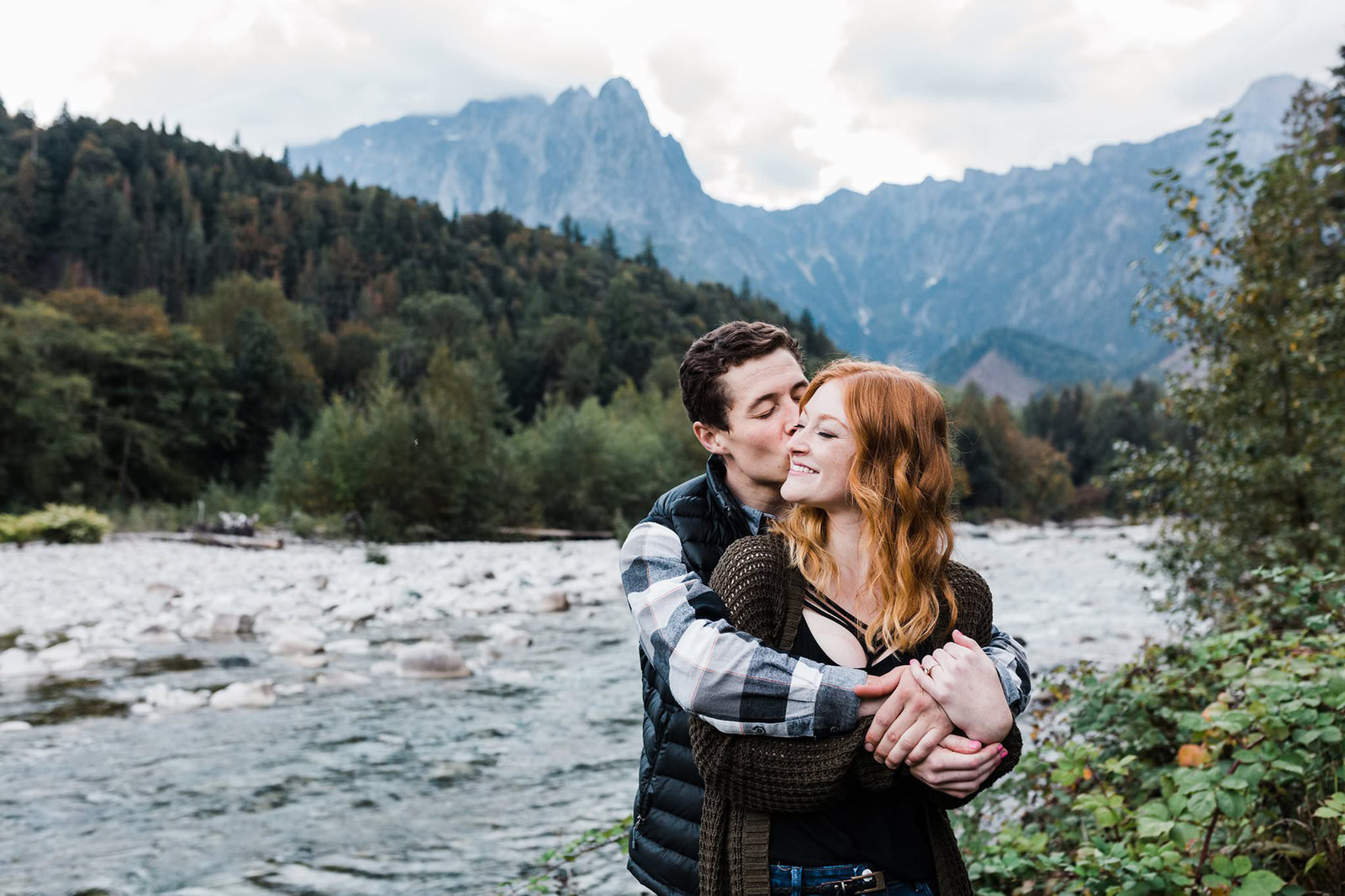 A couple snuggles and stays warm next to a river in Index, Washington during their adventure engagement session in the mountains of Washington