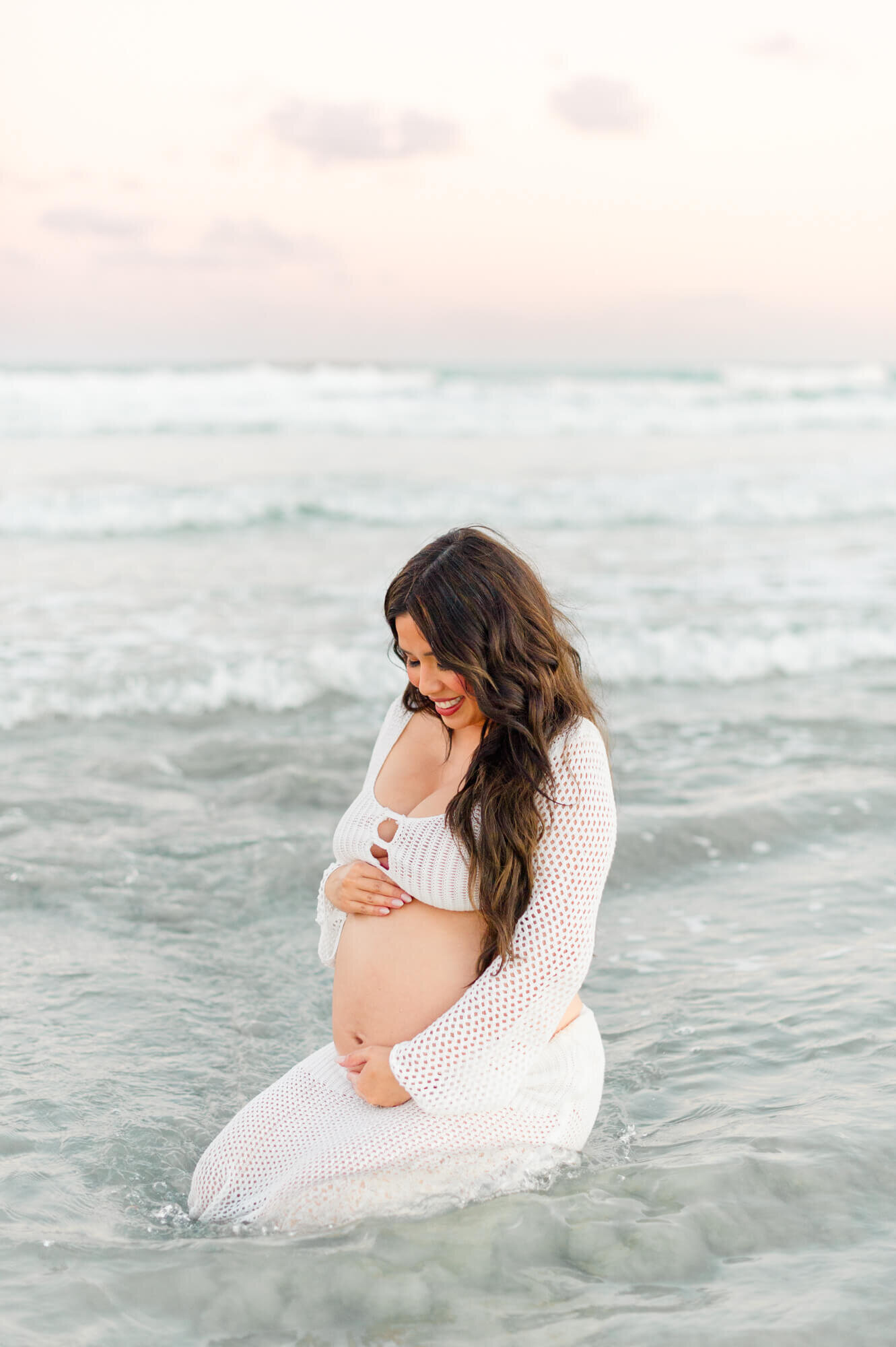 New mother holds her belly in the ocean while looking down at her baby boy