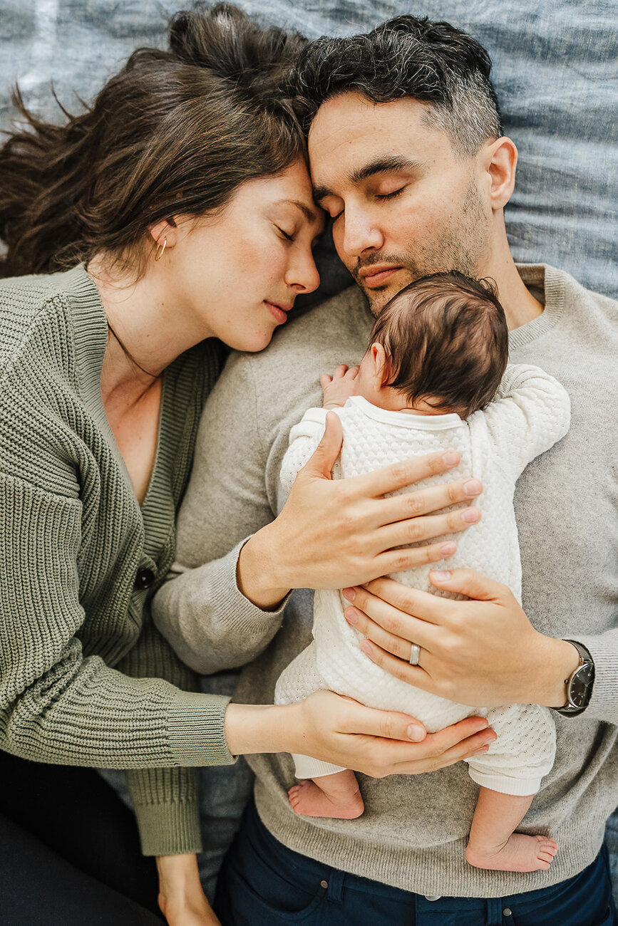 new parents lay on bed snuggling with newborn on chest in boston