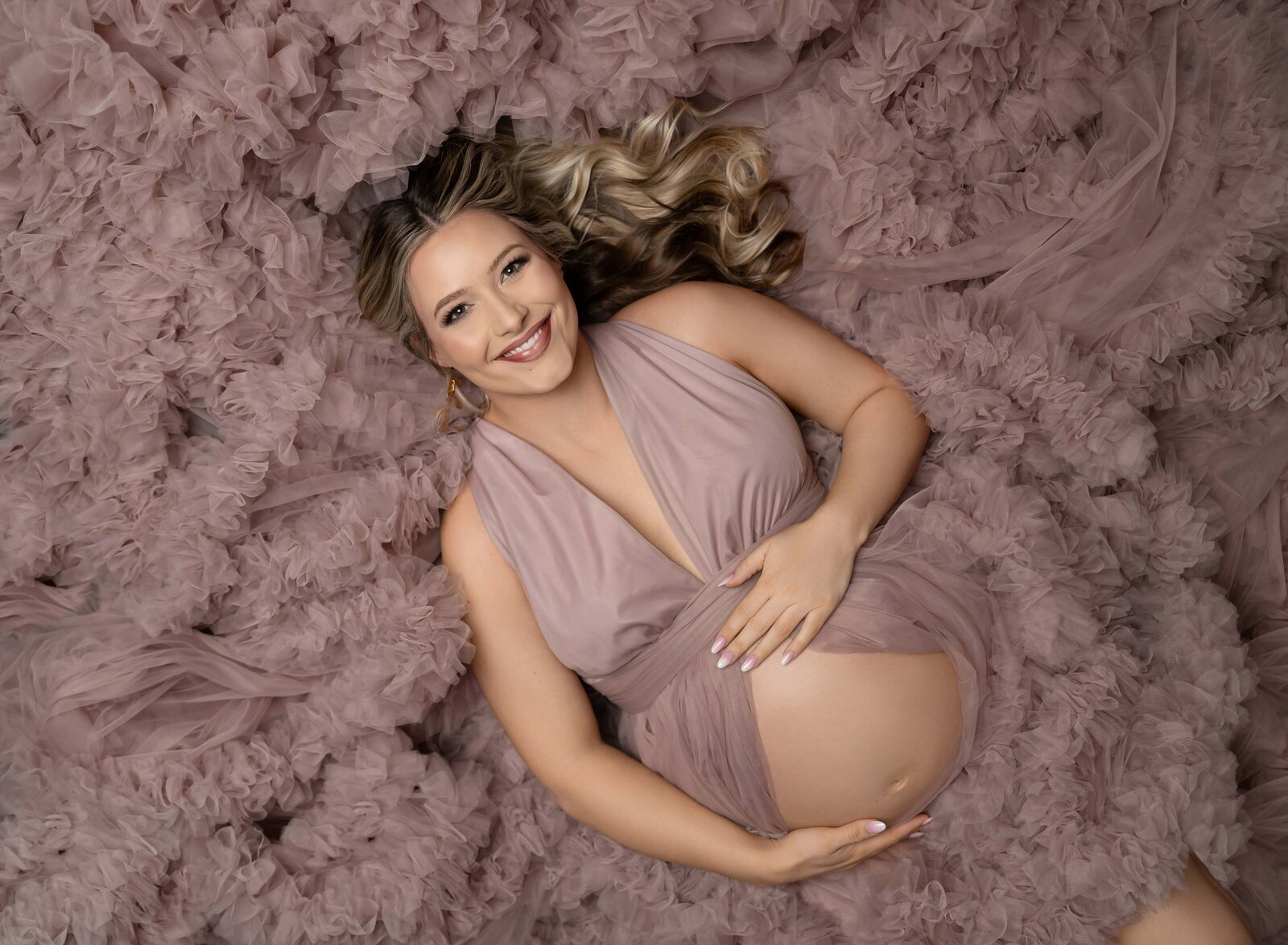 Mom holding baby bump and laying in a bed of pink tulle. Maternity portrait taken by Jenni. Nikki Photography at Atlanta Photo Studio. 