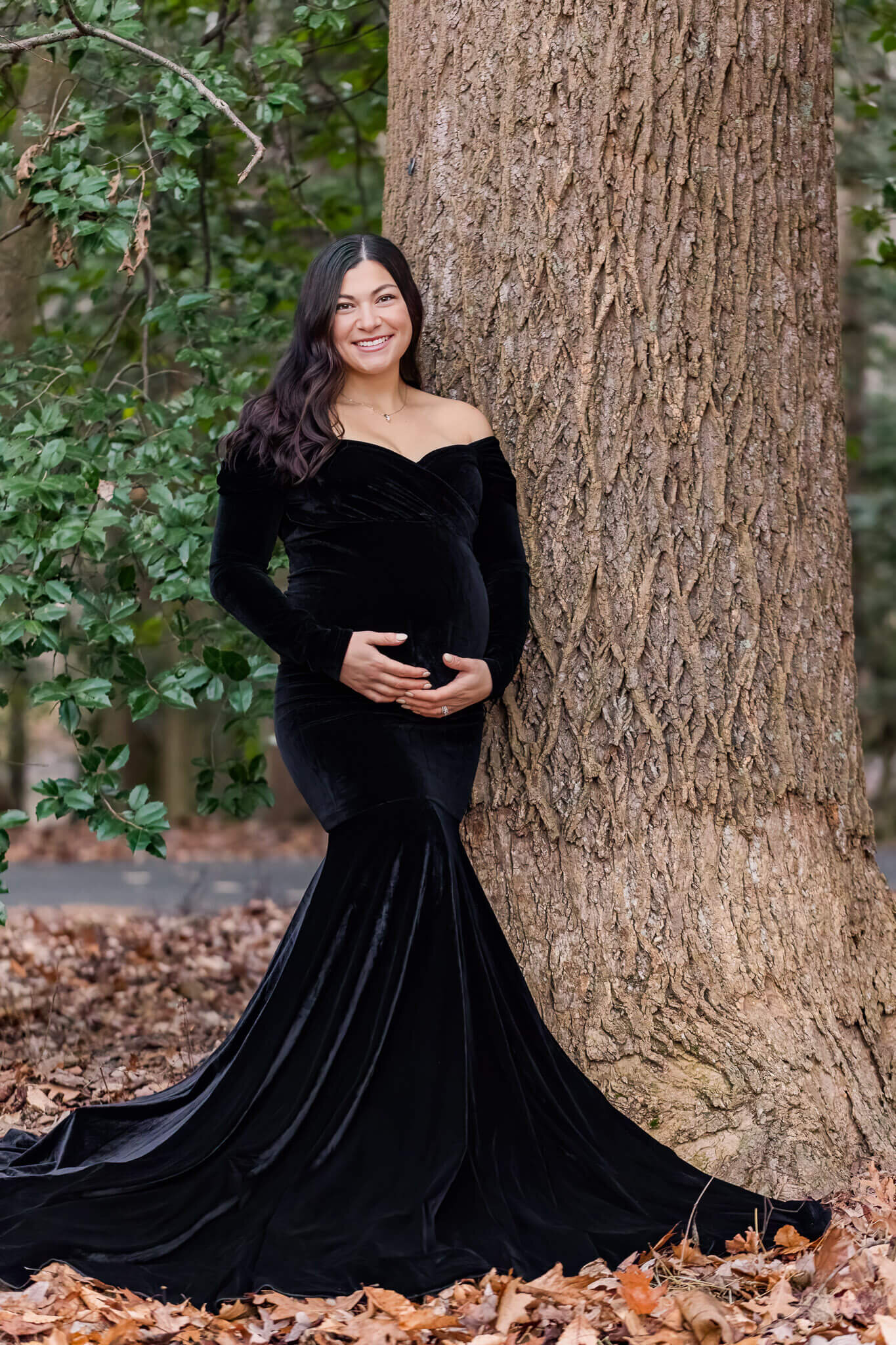 A momma-to-be holding her pregnant belly and leaning against a tree at a park.