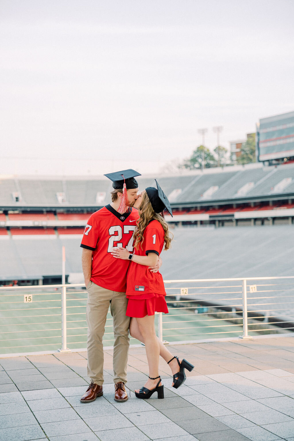 Girl and guy in red UGA shirts and graduation caps kiss in UGA stadium