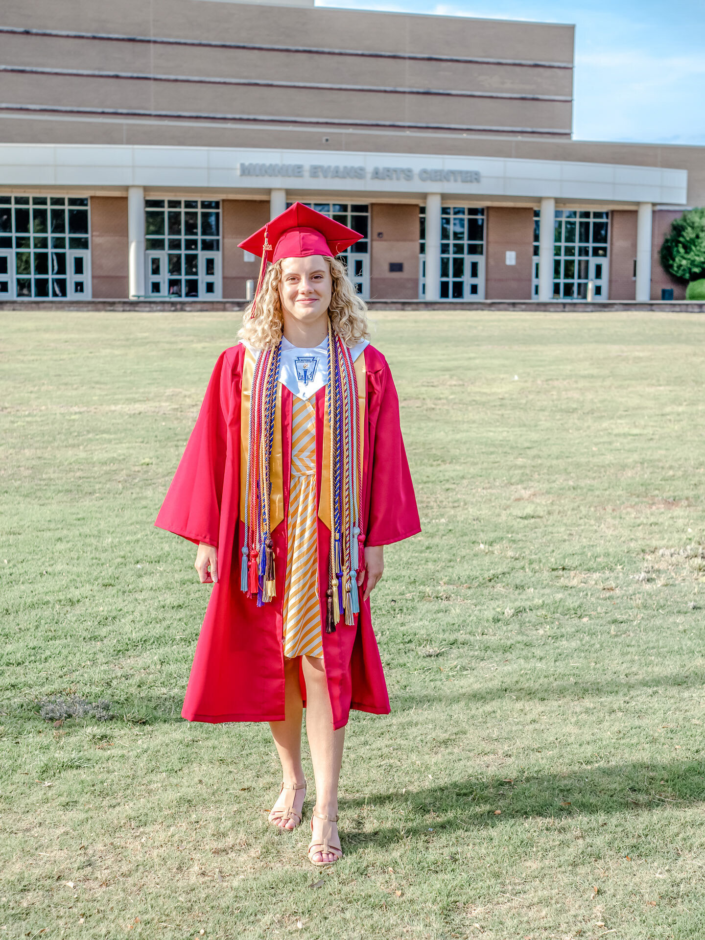 An Ashley High School senior in a red graduation cap and gown with honor cords standing in front of the Minnie Evans Arts Center in Wilmington, North Carolina. This photo celebrates academic achievement, ideal for those seeking professional senior photography.