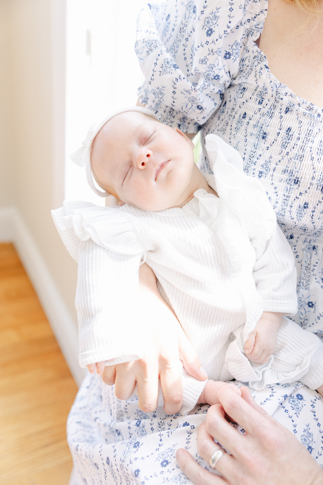 A serene newborn baby sleeps in her mother’s arms, wrapped in a white ruffled outfit and headband. The soft light streaming through the window captures the warmth and tenderness of an in-home newborn session in Connecticut. This image highlights the ease and comfort of documenting those first precious days without ever leaving the house.