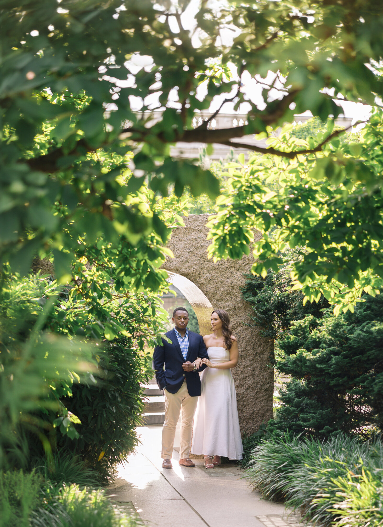 A couple stands together in a lush garden, framed by a stone arch and surrounded by green foliage. The woman wears a white dress, and the man is in a blazer and light pants. Sunlight filters through the leaves, creating a serene atmosphere.