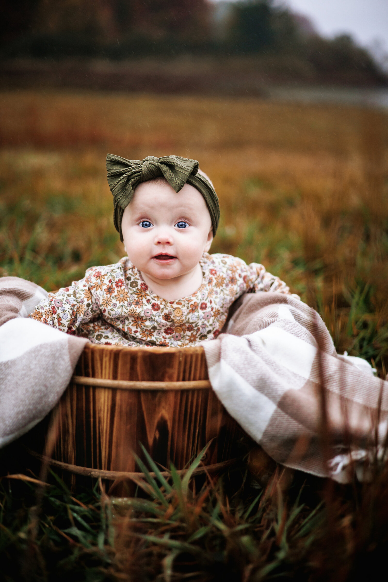 Sweet baby girl sits in a wooden bucket amongst  the grass on a foggy day