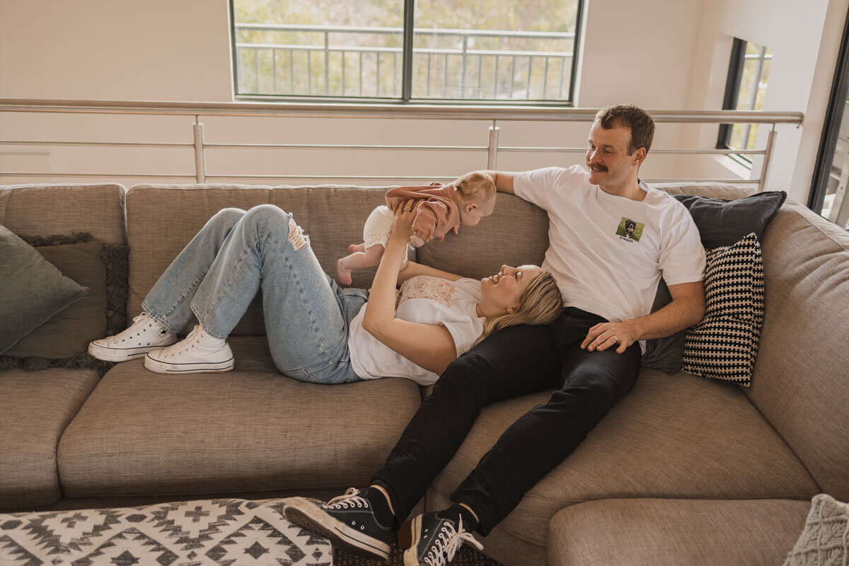 New parents hold their baby up on the couch.