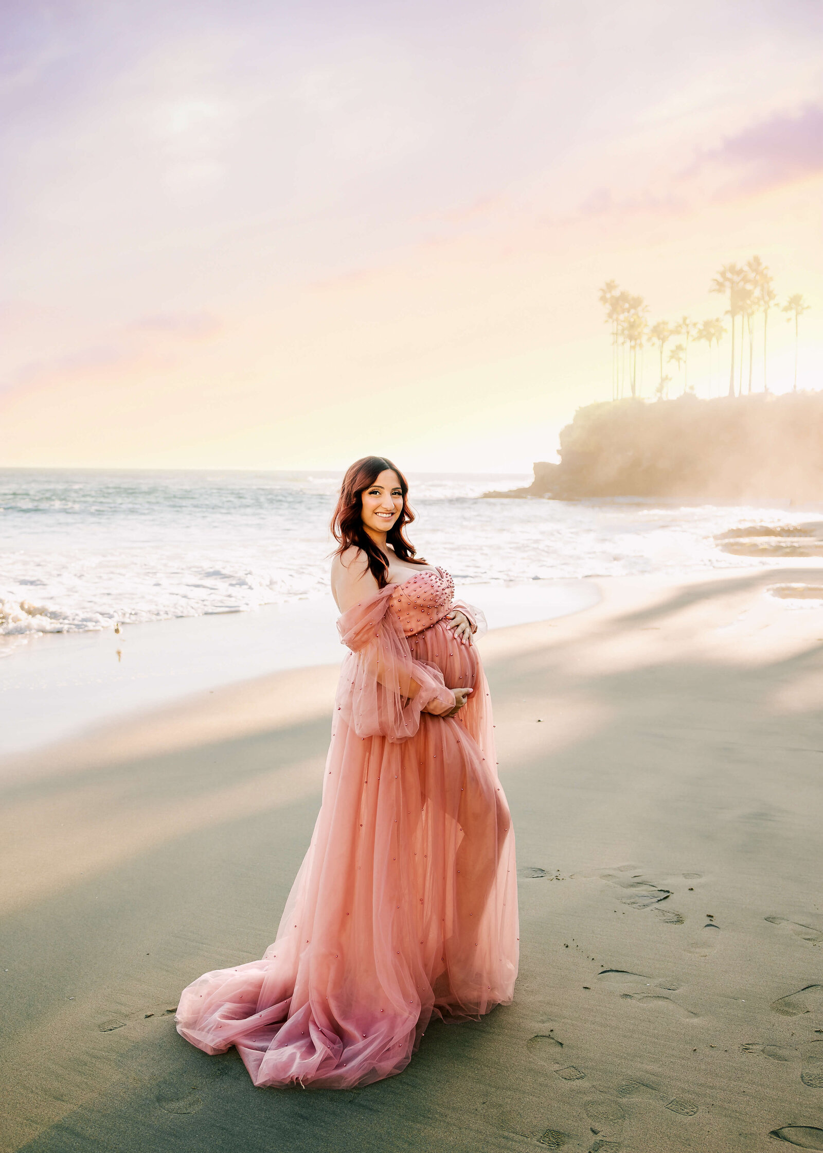 Mom holding belly at laguna beach maternity session wearing pink pearl dress smiling at sunset.