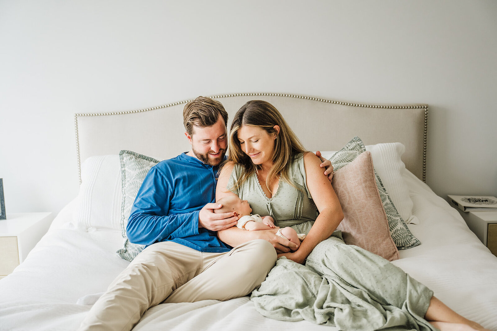 mom and dad sit on neutral bed ad gaze down at their infant son