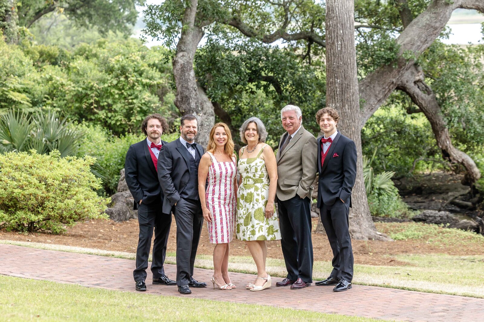 A family poses in a lush garden, dressed in formal attire with men in suits and women in dresses. The greenery and pathways create an elegant atmosphere, ideal for artistic family photography in Wilmington, NC, capturing sophisticated family moments.