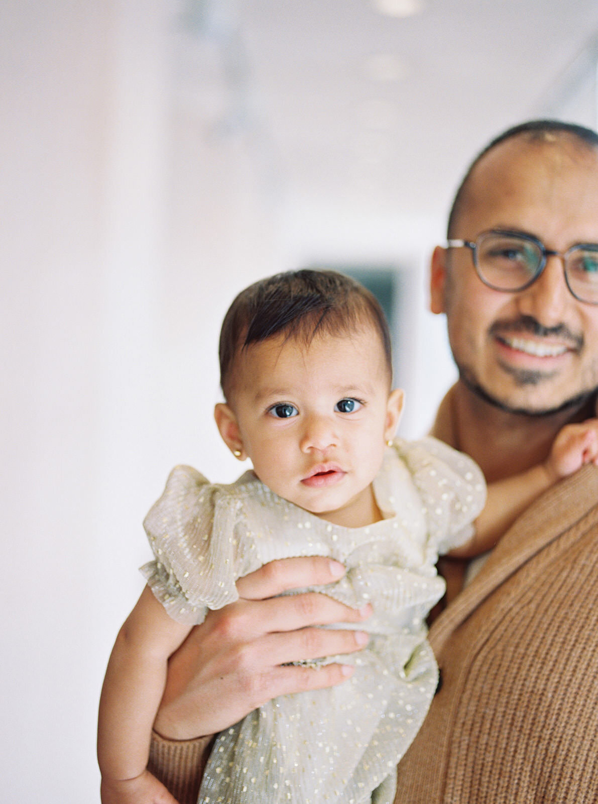 A heartfelt portrait of a father holding his baby daughter, dressed in a shimmering gold dress, as she gazes directly at the camera with wide, curious eyes, exuding elegance and warmth.