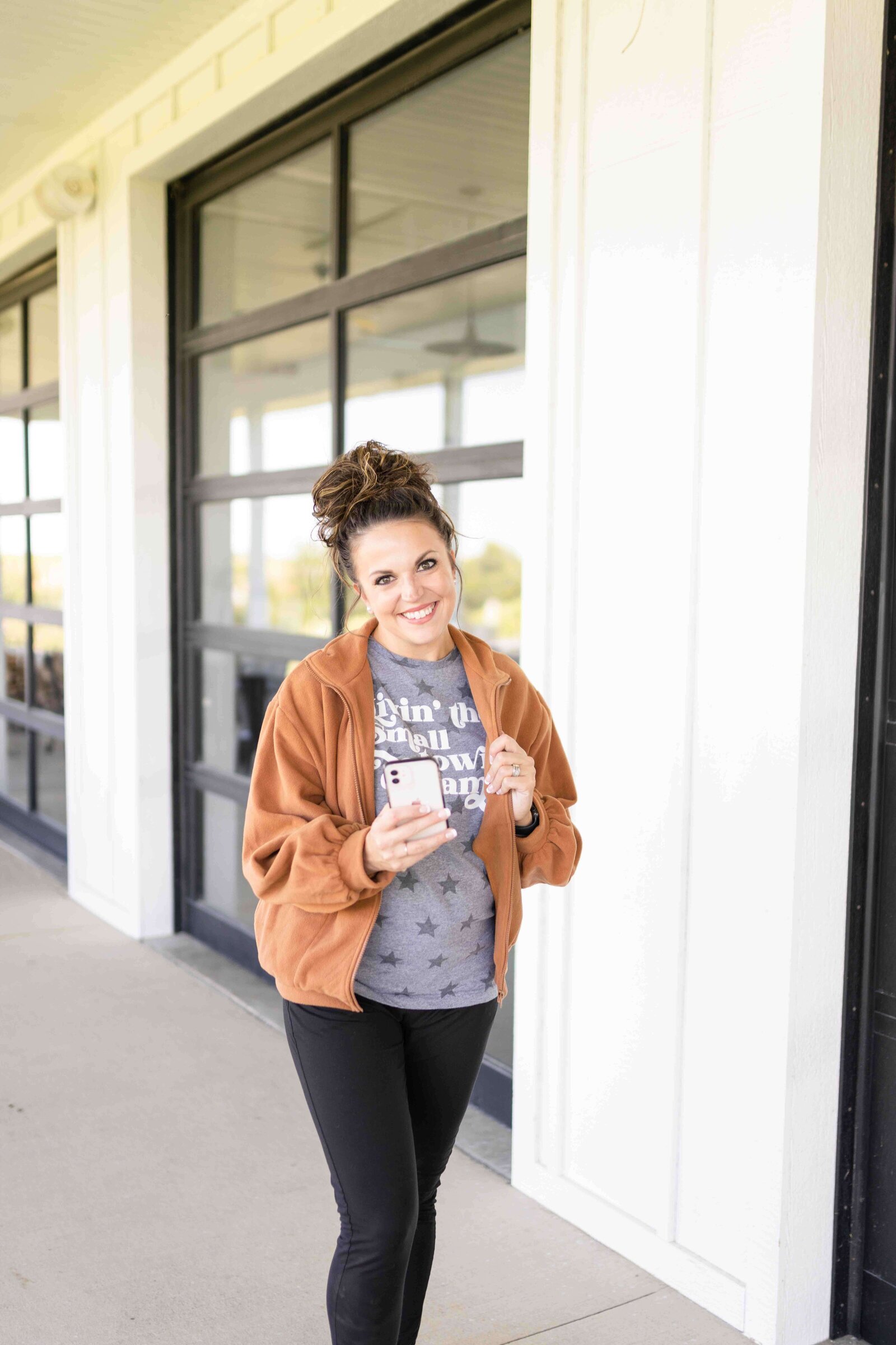Woman smiling outside wearing small town tshirt and brown jacket holding cell phone