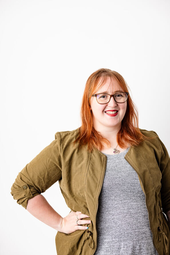 Woman with red hair and glasses in a green jacket, puts her hand on hip and poses on a white background.