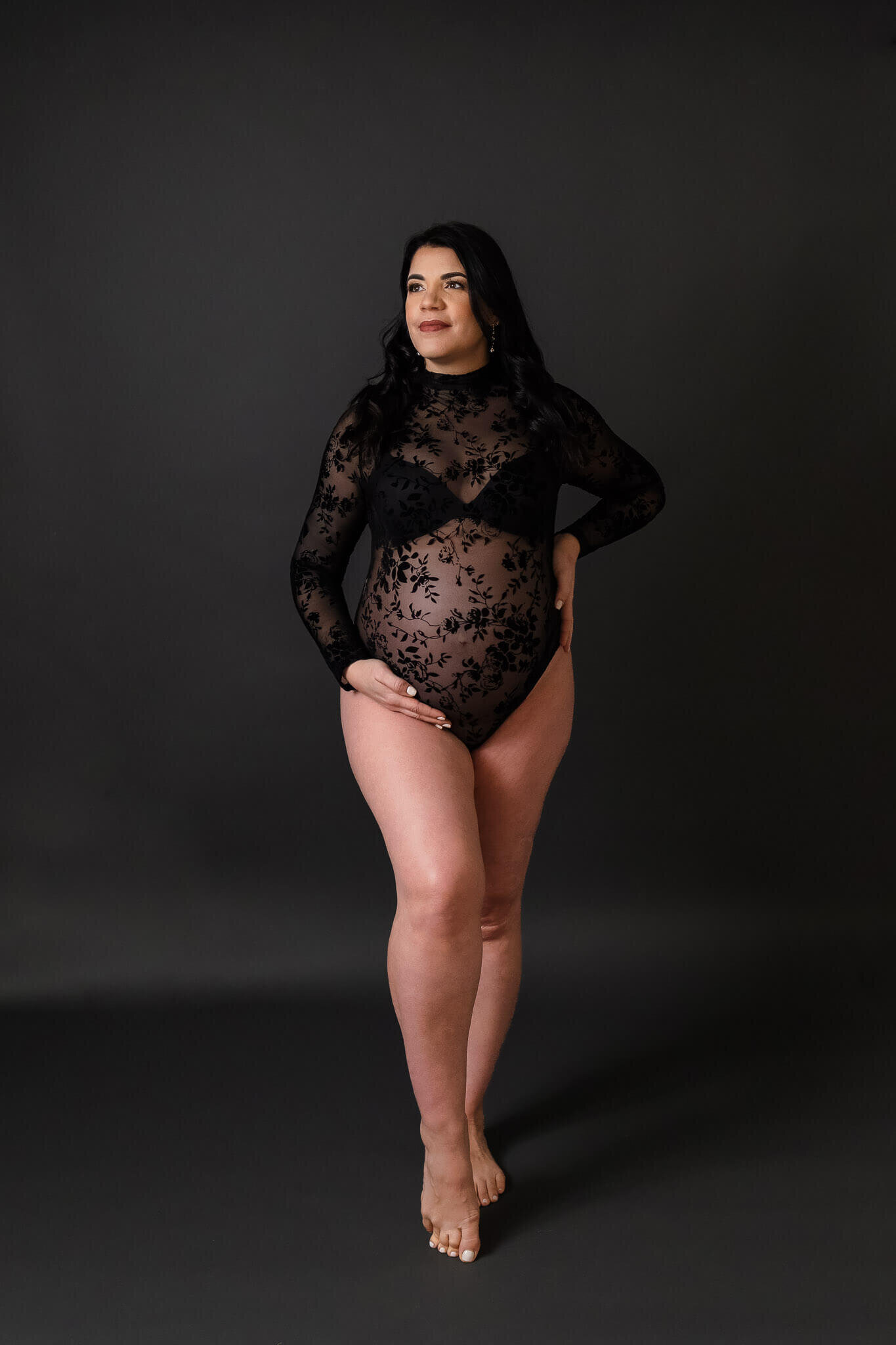 A mom-to-be wearing a lace black bodysuit and posing on a gray backdrop.