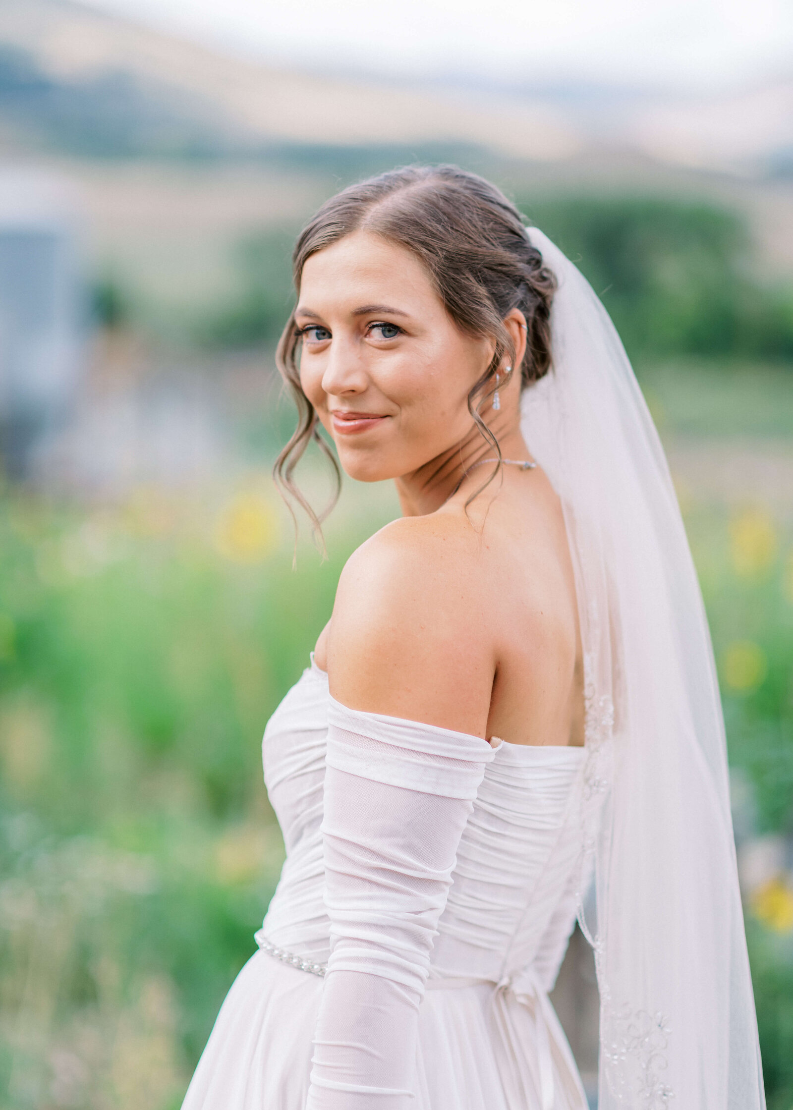 Beautiful bride looks over her shoulder while standing in a green field filed with wildflowers. Image by virginia wedding photographer Erin Winter