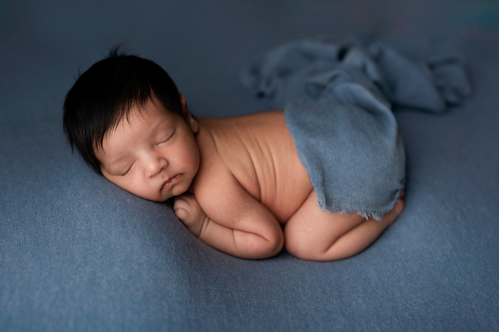 Baby boy with a full head of black hair, posed on his belly in front of a blue back drop.