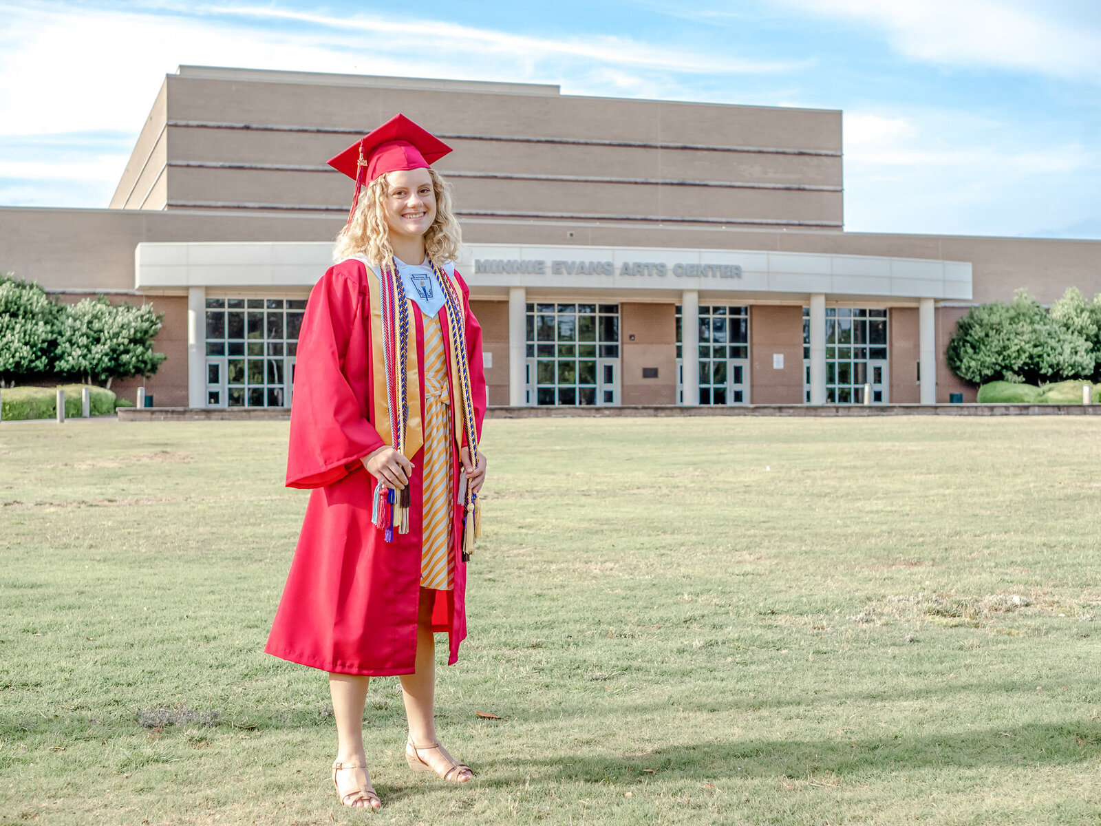 A Ashley High School senior in a red graduation gown and honor cords standing in front of the Minnie Evans Arts Center in Wilmington, North Carolina. This photo celebrates academic achievement, perfect for capturing memorable senior moments.