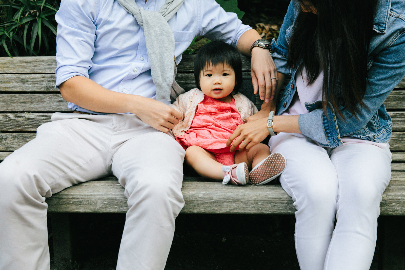 Chinese baby girl in pink dress sitting on a park bench between her parents at San Francisco Botanical Gardens