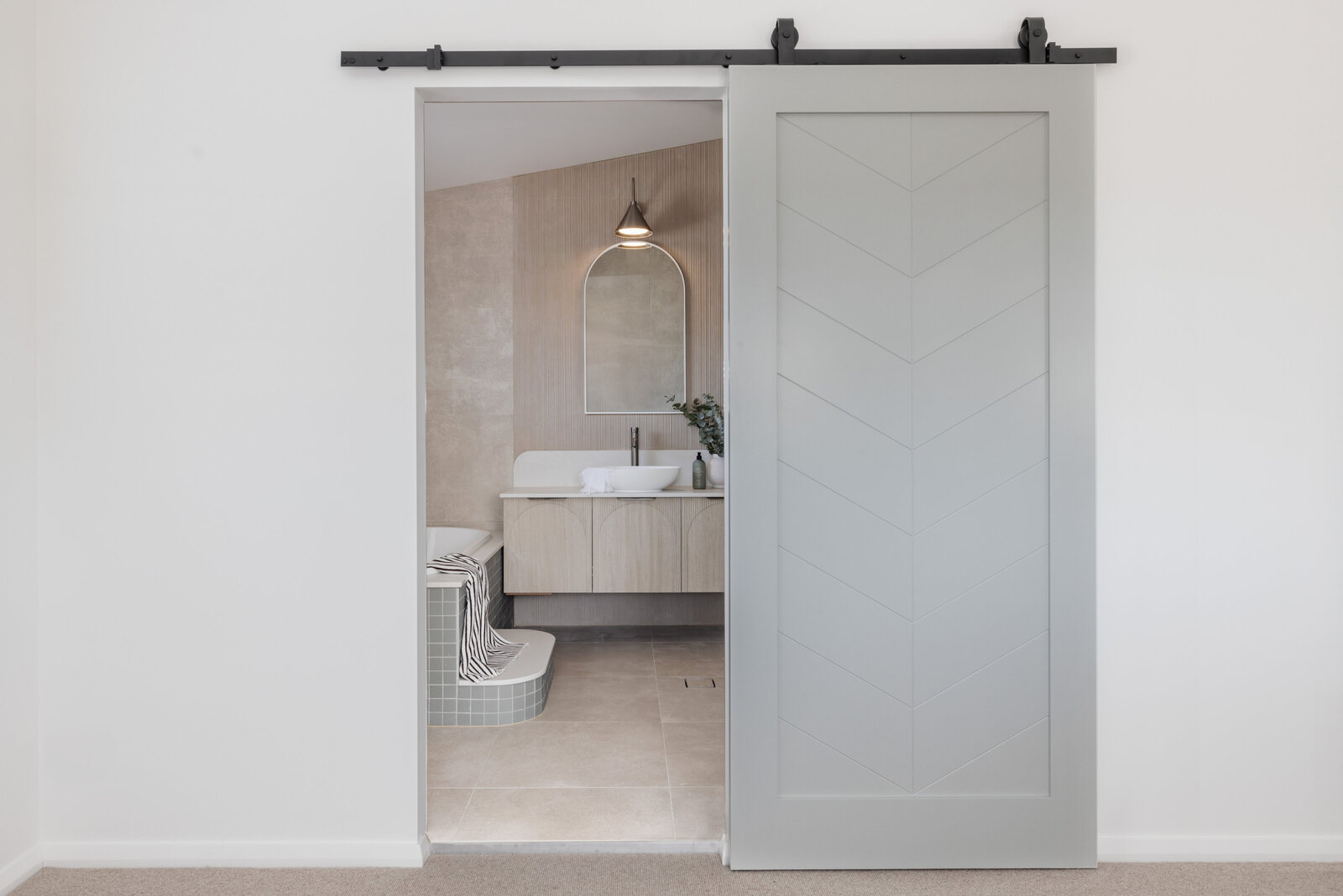 Sliding barn door opening to a modern bathroom with a wall-mounted sink, circular mirror, and neutral tones by Newcastle Kitchen and Bathroom Designer.