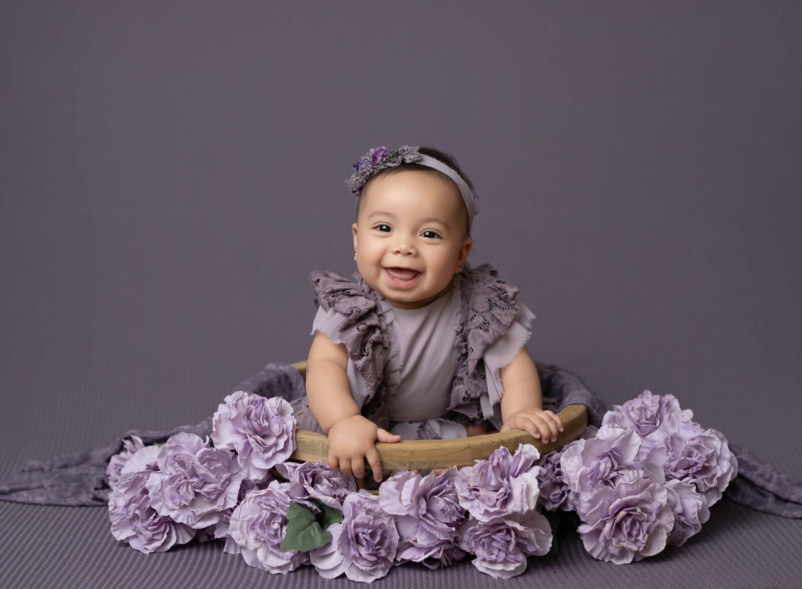 Baby girl in purple romper sitting in a wooden basket surrounded by purple flowers. Photographed at Atlanta baby photo studio.