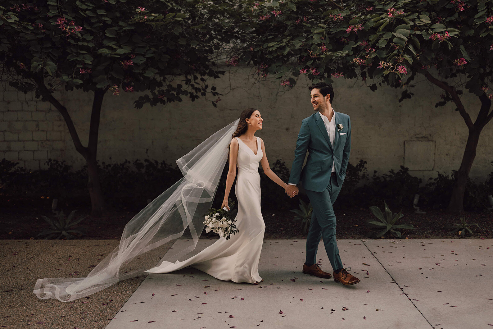 A bride and groom walk hand-in-hand beneath flowering trees outside SoHo63, a Chandler wedding and event venue. The bride’s long veil flows behind her as the couple smiles at each other, surrounded by soft natural light and lush greenery.