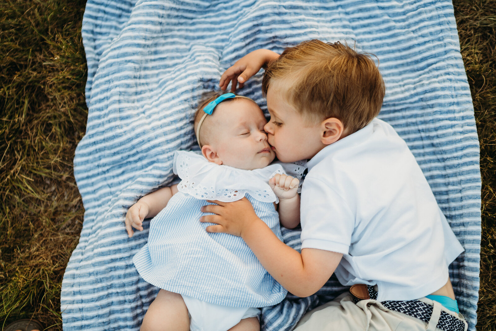 big brother kisses baby sister on a blue striped blanket
