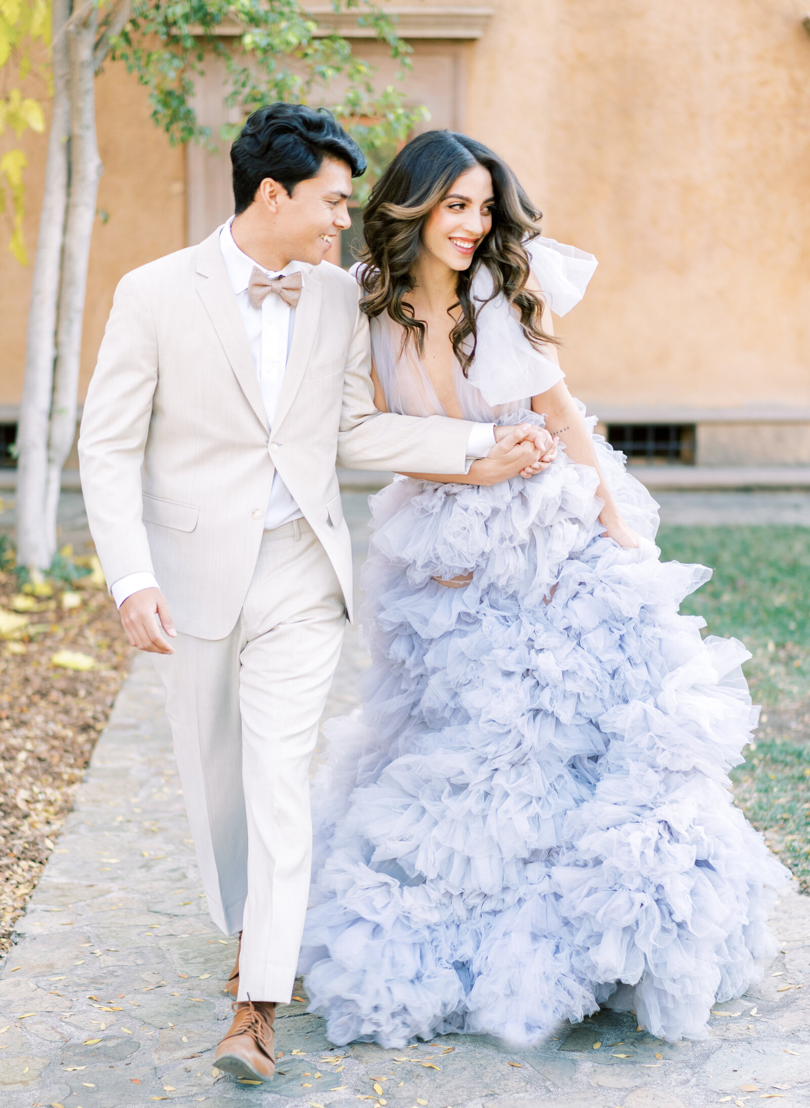 Portrait of bride and groom in a lavender gown and cream suit walking outdoors by a peach-colored structure.